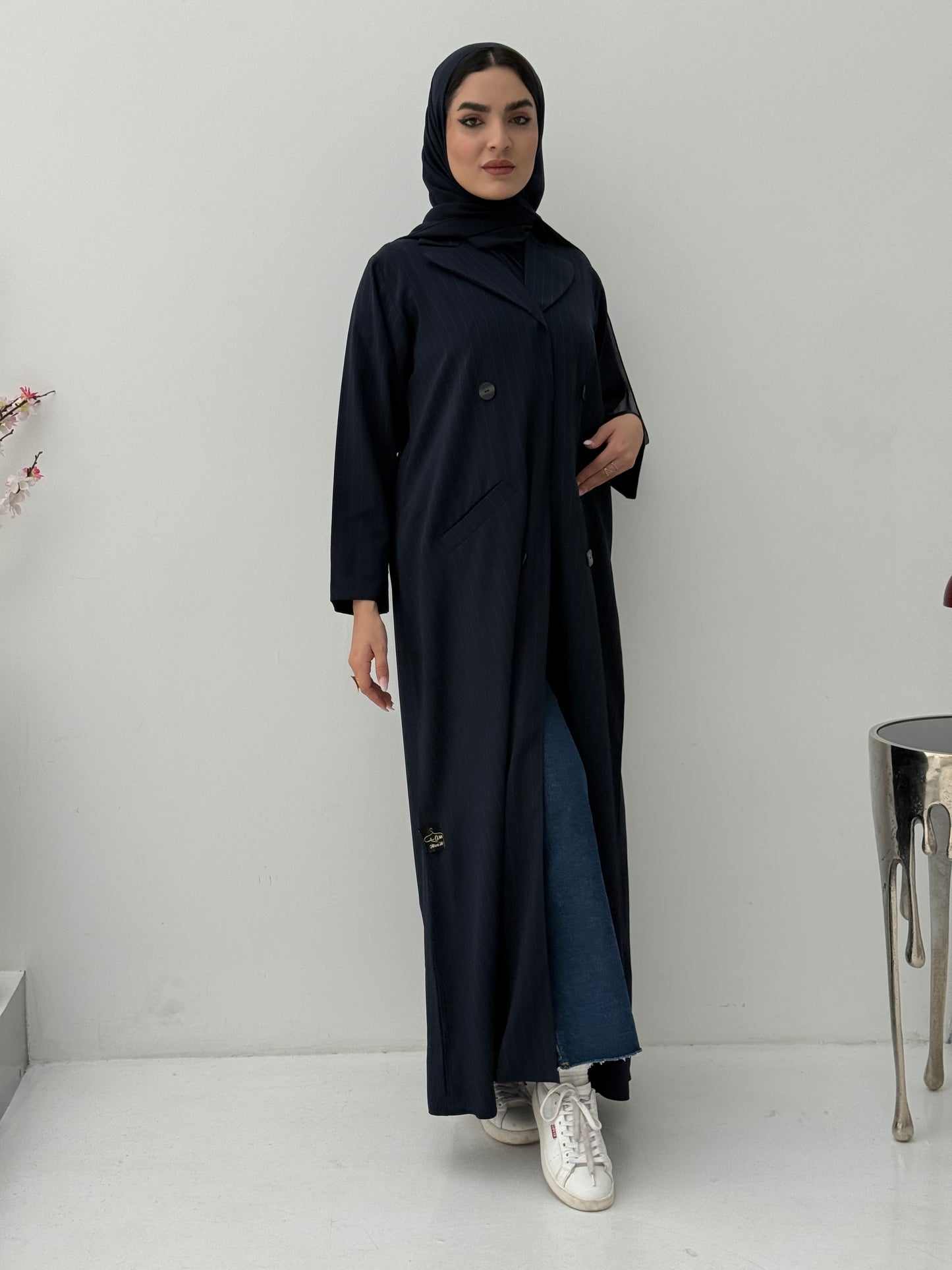 A woman wearing a navy blue pinstripe linen coat, with a sheila on her head, standing against a white background.