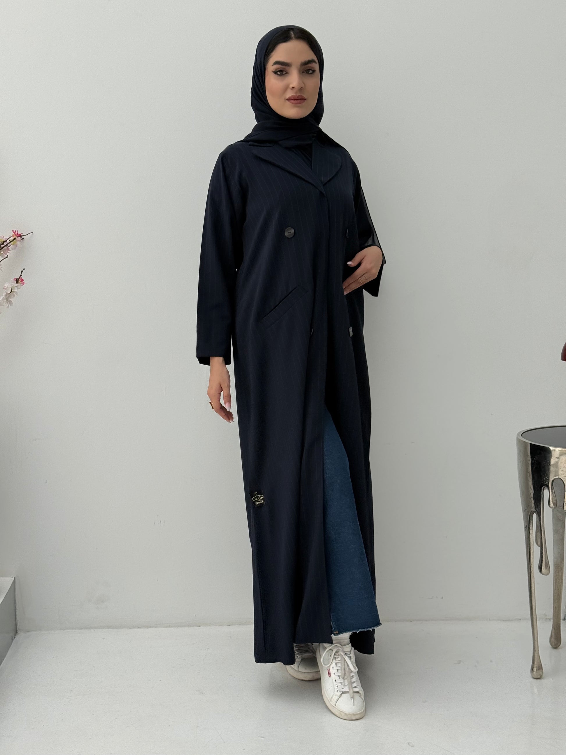 A woman wearing a navy blue pinstripe linen coat, with a sheila on her head, standing against a white background.