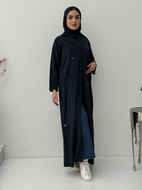 A woman wearing a navy blue pinstripe linen coat, with a sheila on her head, standing against a white background.