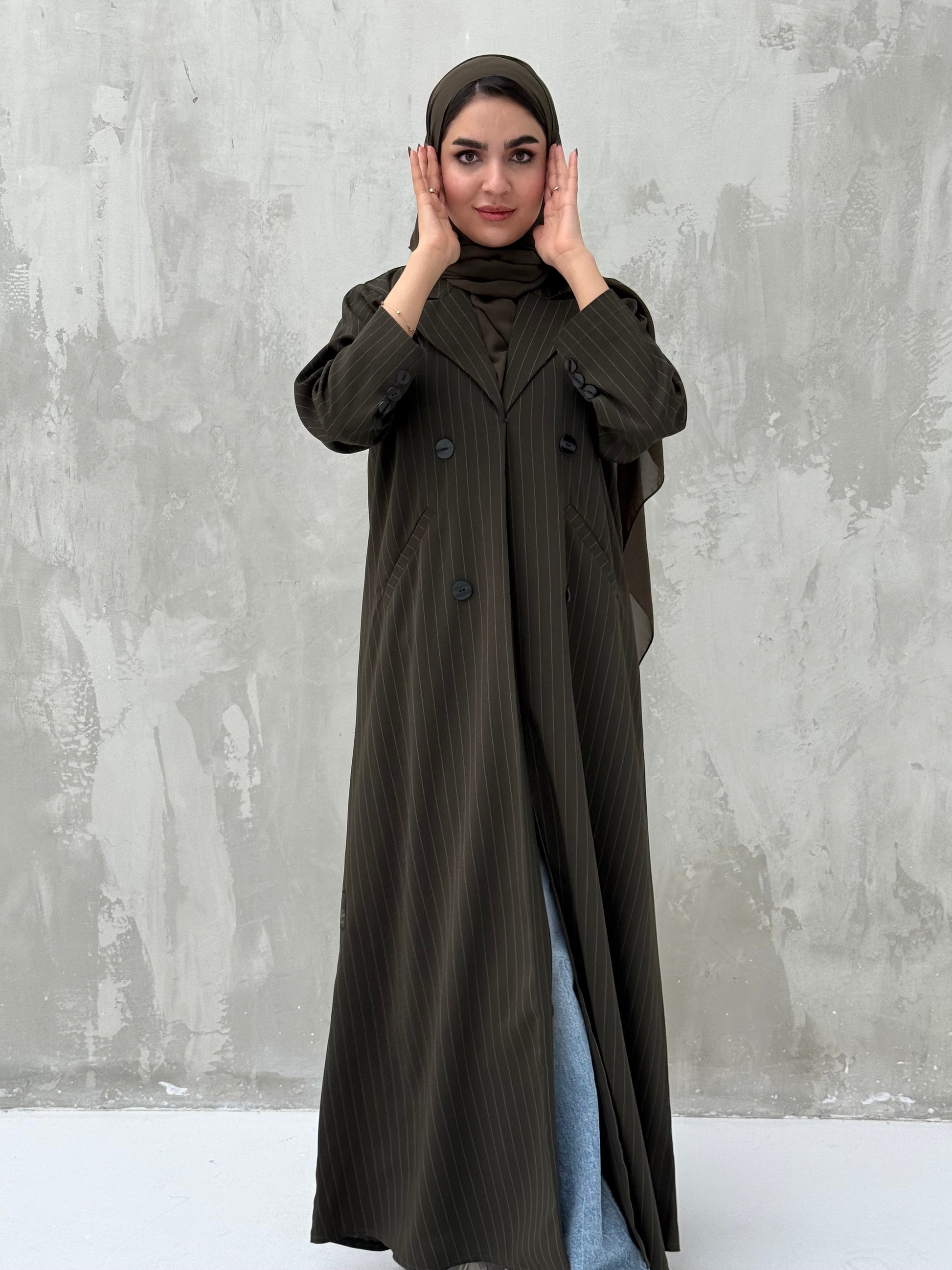 A woman wearing an olive green pinstripe linen coat, with a sheila on her head, standing against a grey concrete background.