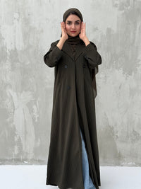 A woman wearing an olive green pinstripe linen coat, with a sheila on her head, standing against a grey concrete background.