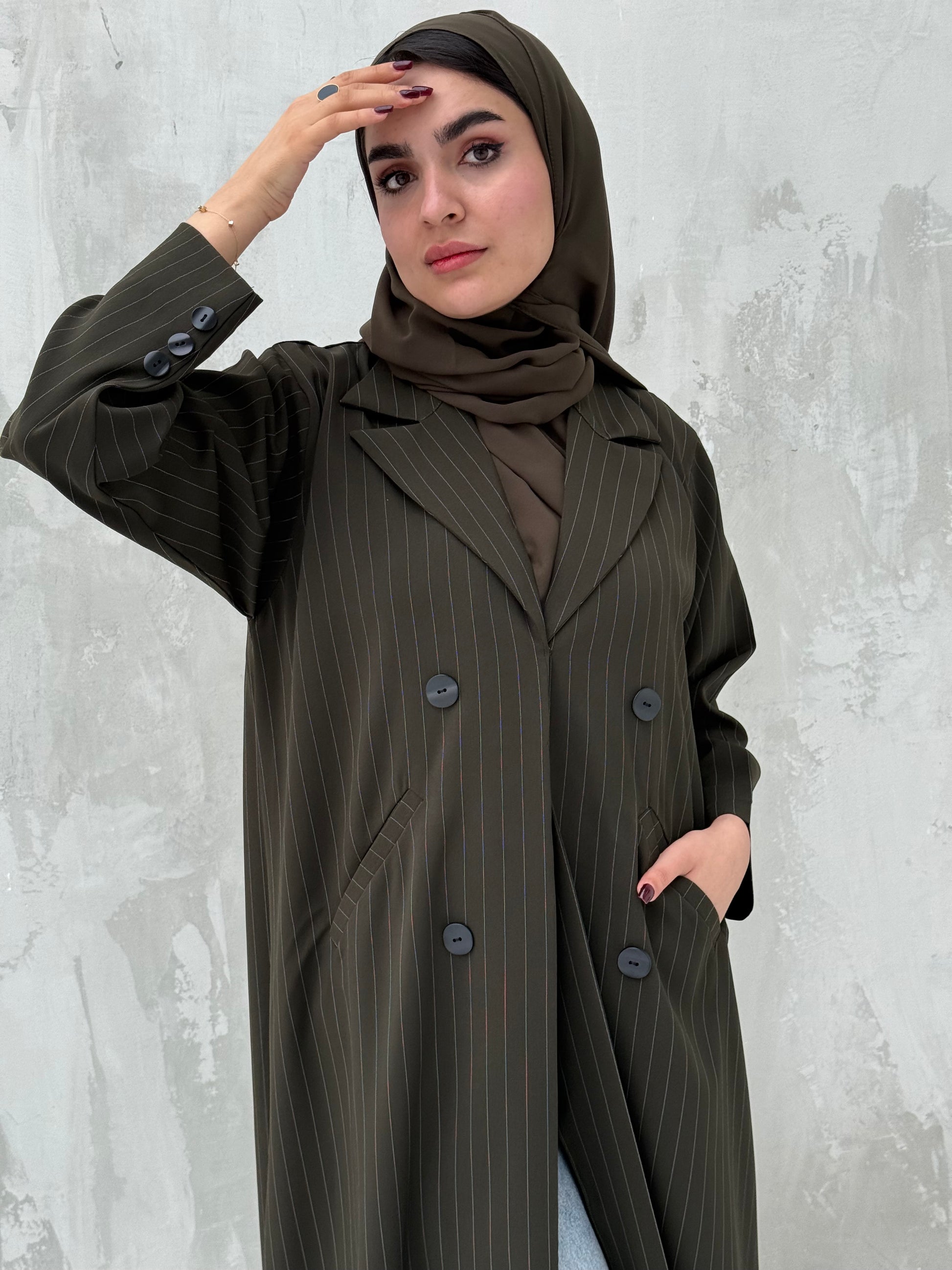 A woman wearing an olive green abaya with a striped pattern, featuring a matching sheila and button-down front.