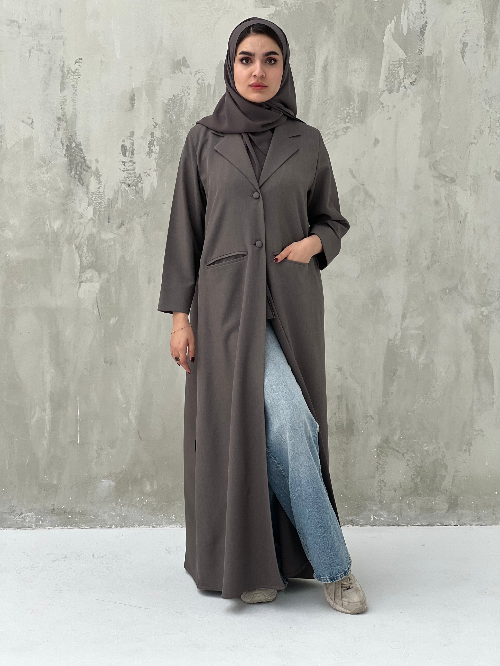 A woman wearing an Ash Brown abaya with a beige sheila, over a black outfit, with blue jeans and beige shoes, standing against a gray background.