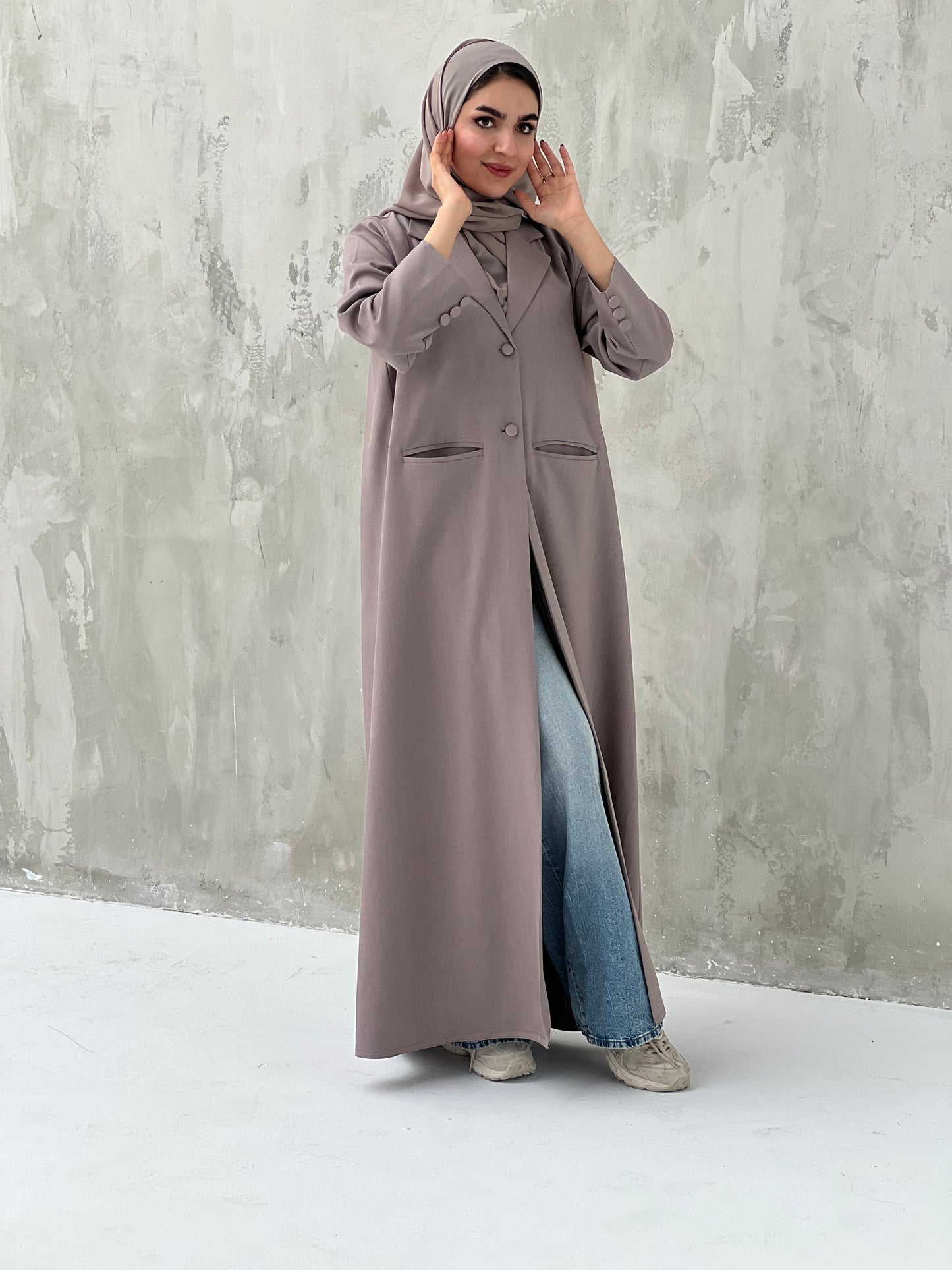 A woman wearing a dark beige abaya with a beige sheila, over a black outfit, with blue jeans and beige shoes, standing against a gray background.