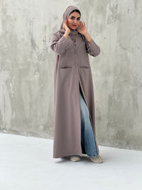 A woman wearing a dark beige abaya with a beige sheila, over a black outfit, with blue jeans and beige shoes, standing against a gray background.