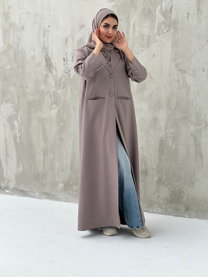 A woman wearing a dark beige abaya with a beige sheila, over a black outfit, with blue jeans and beige shoes, standing against a gray background.