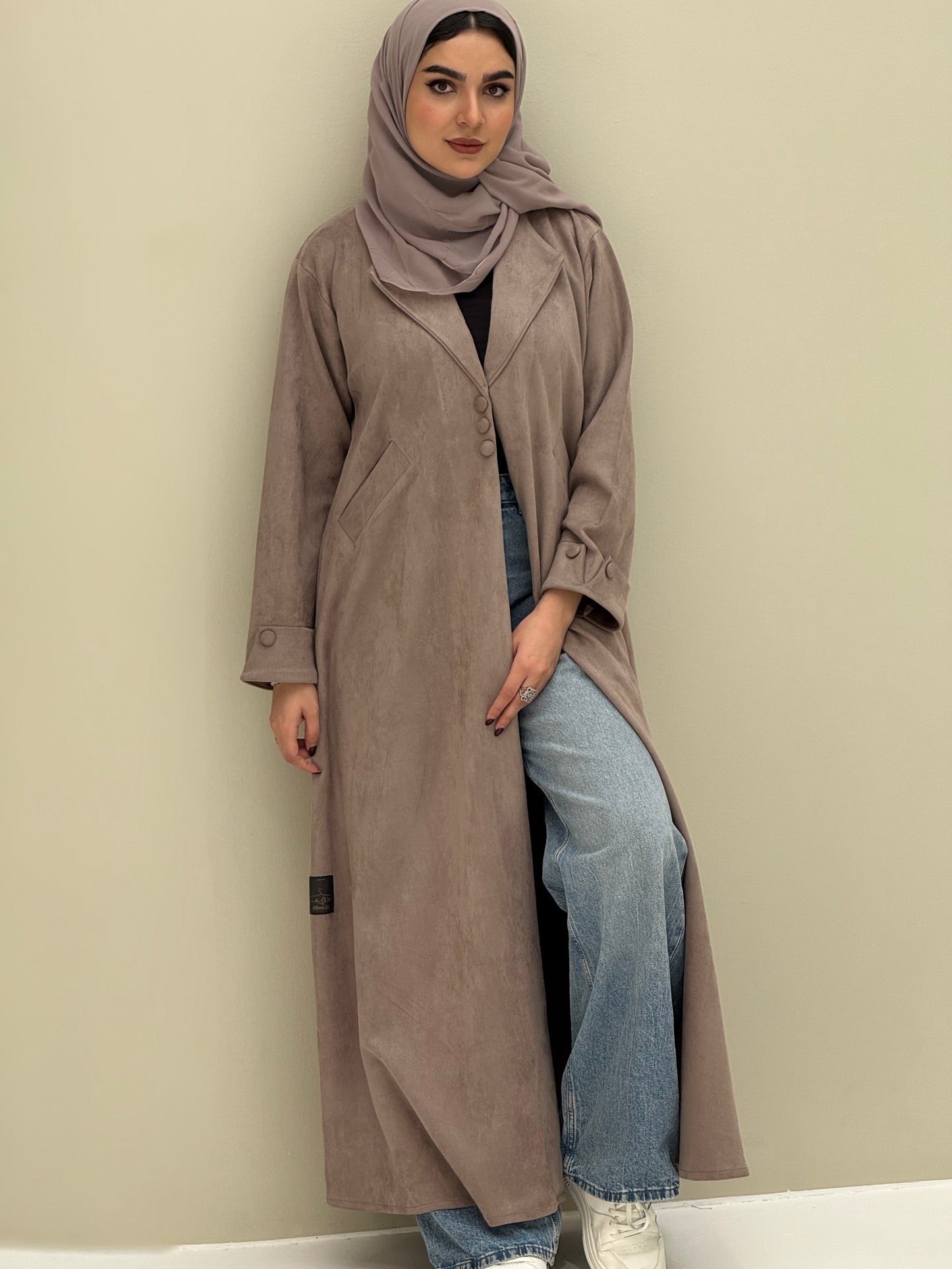 A woman wearing a beige shamwa Coat style abaya with lapel collar, Button accents on front, angled pockets and matching sheila on a cream background