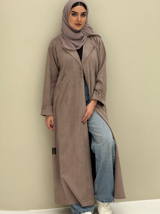 A woman wearing a beige shamwa Coat style abaya with lapel collar, Button accents on front, angled pockets and matching sheila on a cream background