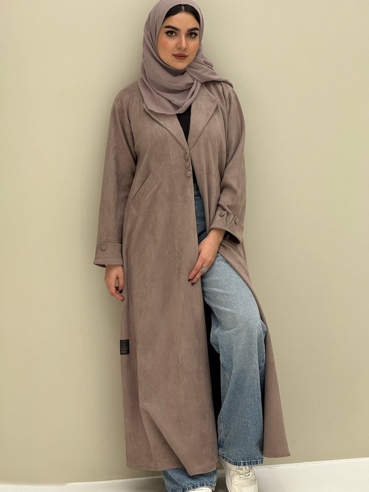 A woman wearing a beige shamwa Coat style abaya with lapel collar, Button accents on front, angled pockets and matching sheila on a cream background