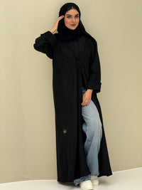 A woman wearing a black shamwa Coat style abaya with lapel collar, Button accents on front, angled pockets and matching sheila on a cream background