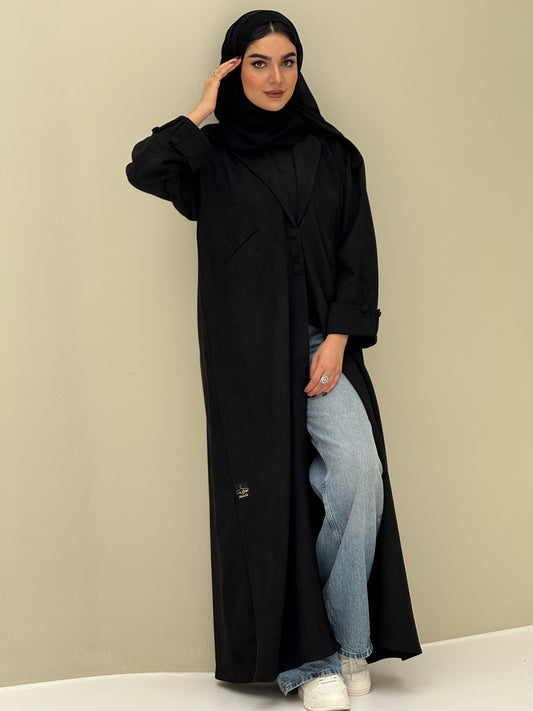 A woman wearing a black shamwa Coat style abaya with lapel collar, Button accents on front, angled pockets and matching sheila on a cream background