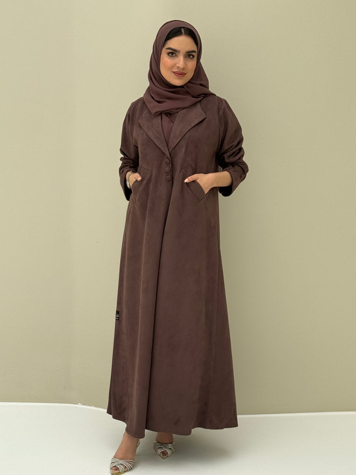 A woman wearing a dark brown shamwa Coat style abaya with lapel collar, Button accents on front, angled pockets and matching sheila on a cream background