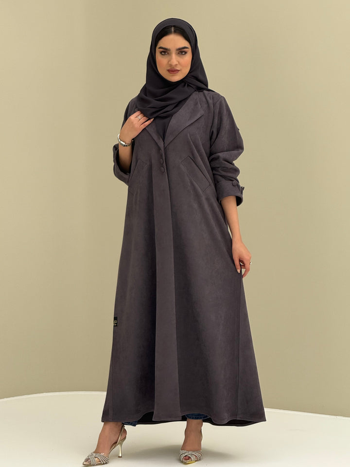 A woman wearing a dark grey shamwa Coat style abaya with lapel collar, Button accents on front, angled pockets and matching sheila on a cream background