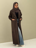 A woman wearing a dark brown abaya with double-breasted buttons, buttoned cuffs, a chest pocket, and a matching sheila paired with blue jeans and white shoes.