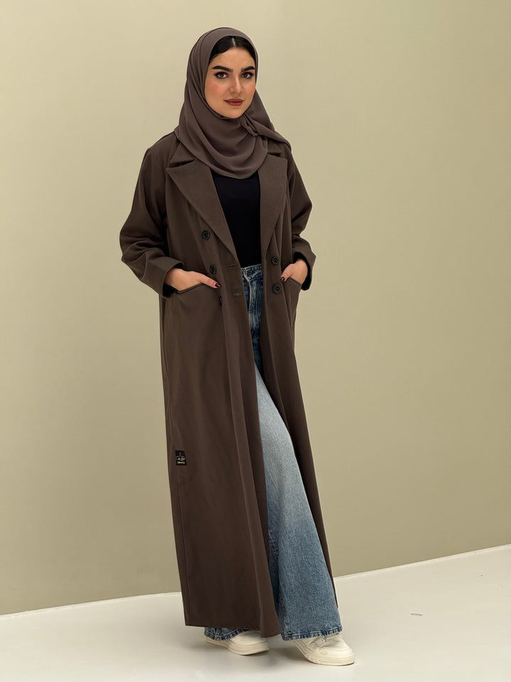 A woman wearing a dark brown abaya with double-breasted buttons, buttoned cuffs, a chest pocket, and a matching sheila paired with blue jeans and white shoes.