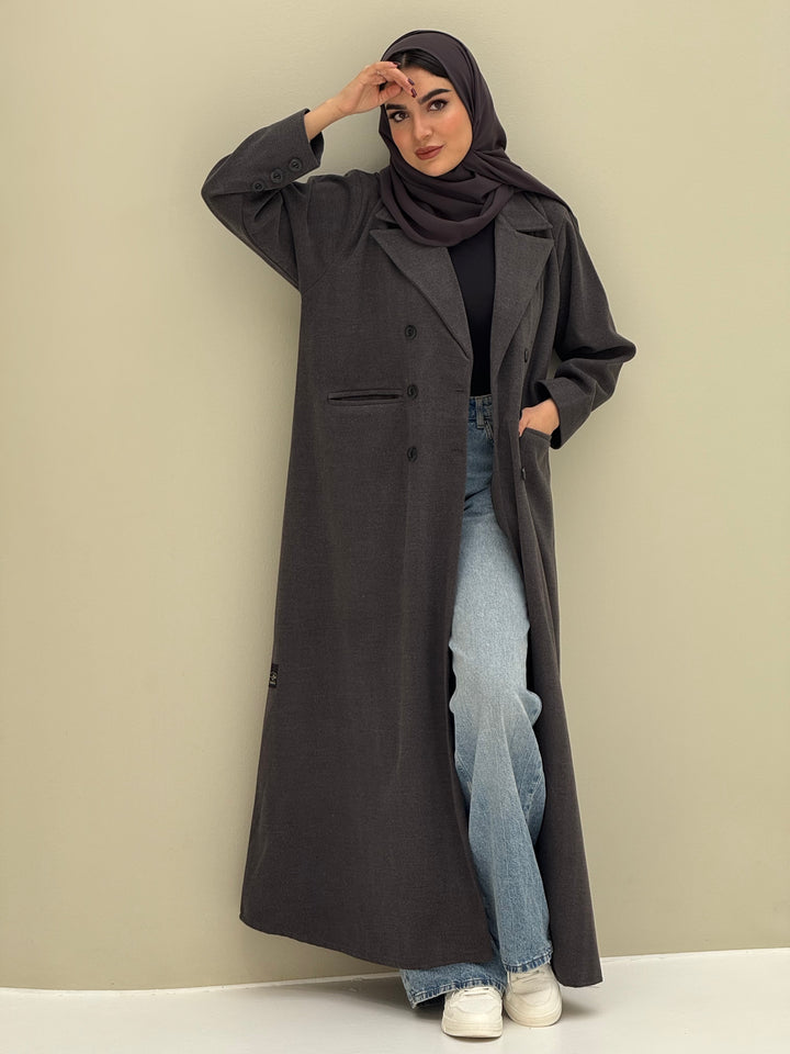 A woman wearing a dark grey abaya with double-breasted buttons, buttoned cuffs, a chest pocket, and a matching sheila paired with blue jeans and white shoes.