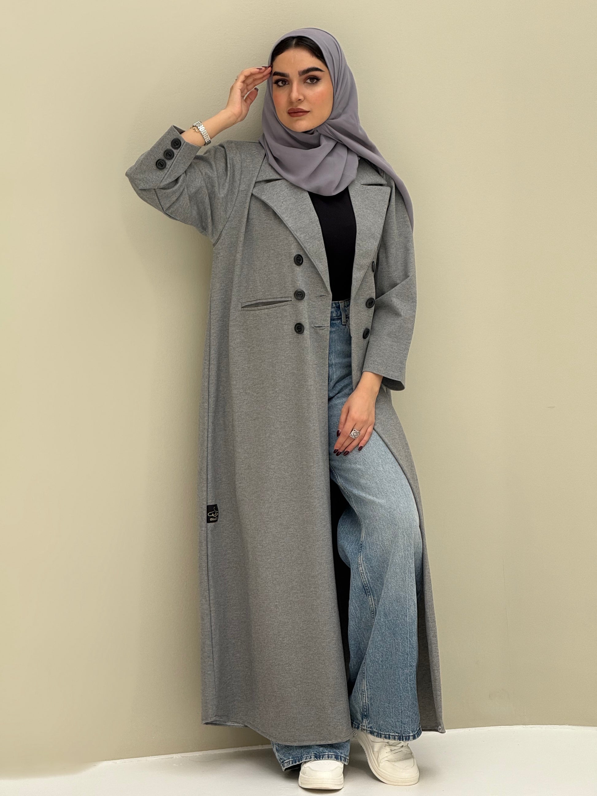A woman wearing a light grey abaya with double-breasted buttons, buttoned cuffs, a chest pocket, and a matching sheila paired with blue jeans and white shoes.