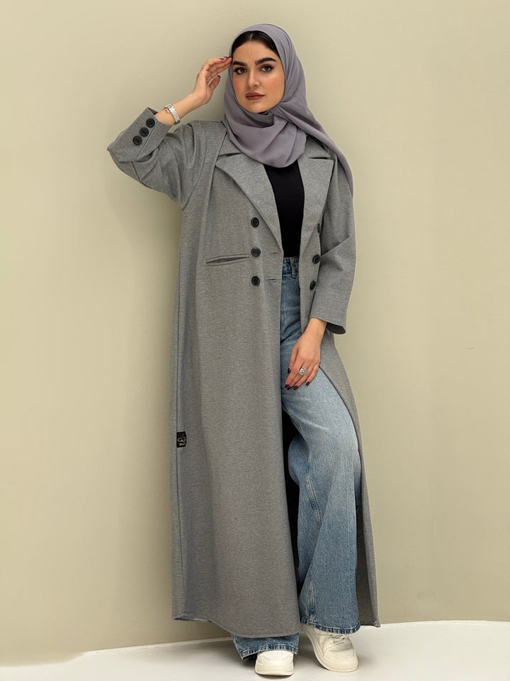 A woman wearing a light grey abaya with double-breasted buttons, buttoned cuffs, a chest pocket, and a matching sheila paired with blue jeans and white shoes.