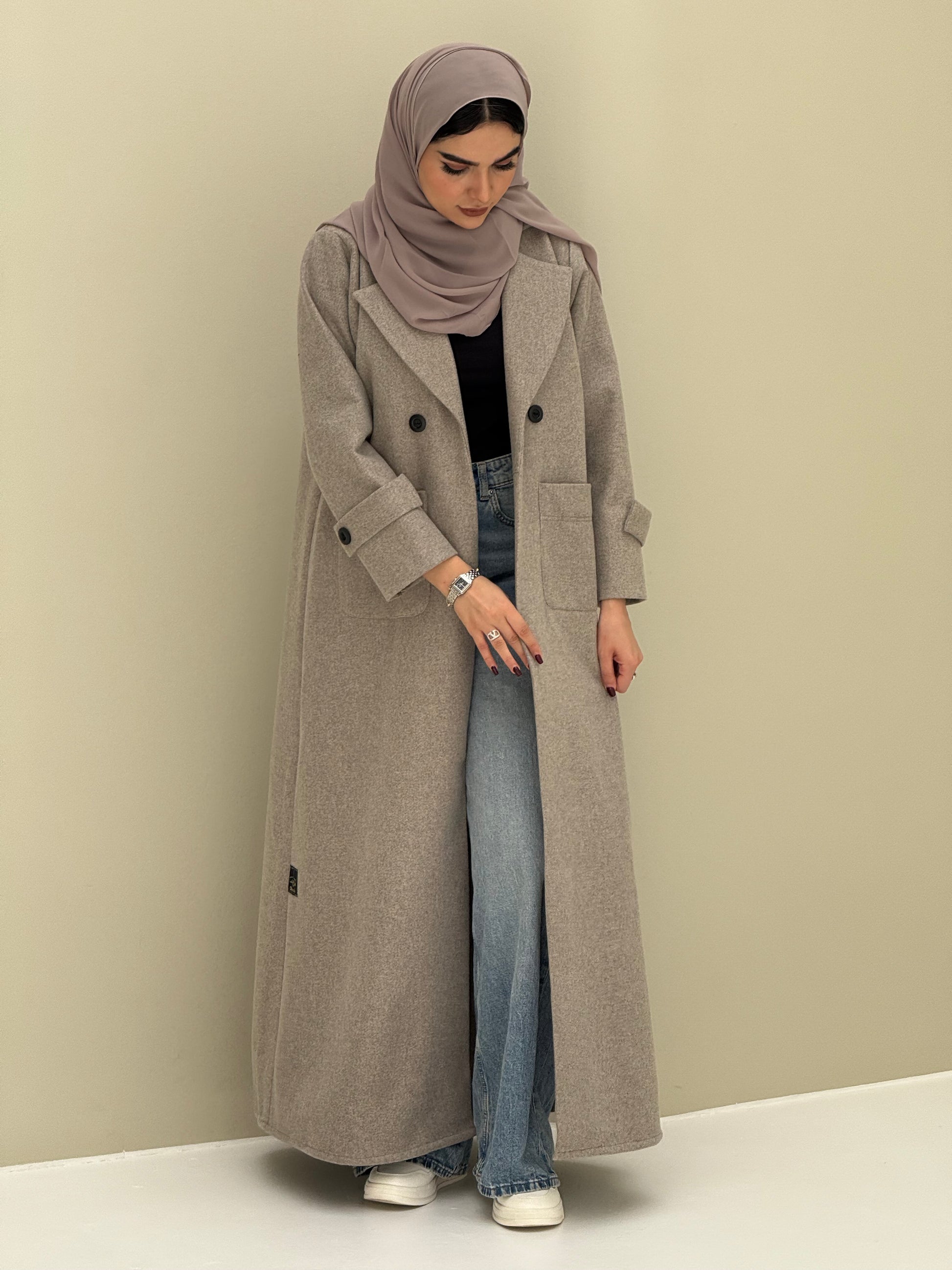 a woman wearing a beige Coat style abaya with lapel collar, Contrast black buttons, large patch pockets, cuff straps and matching sheila