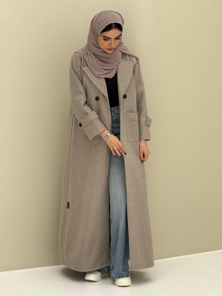 a woman wearing a beige Coat style abaya with lapel collar, Contrast black buttons, large patch pockets, cuff straps and matching sheila