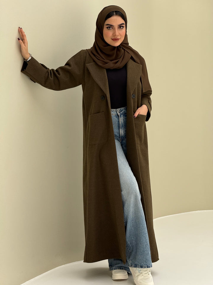a woman wearing a olive green Coat style abaya with lapel collar, Contrast black buttons, large patch pockets, cuff straps and matching sheila