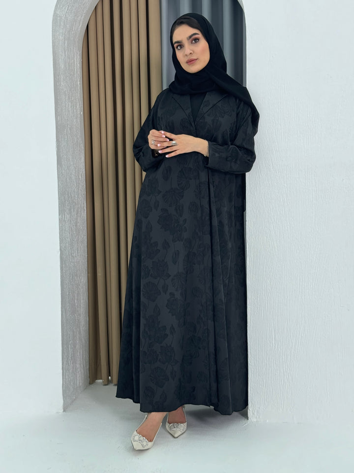 A woman wearing a black abaya with a jacquard fabric, including a matching sheila headscarf, standing against a beige background.