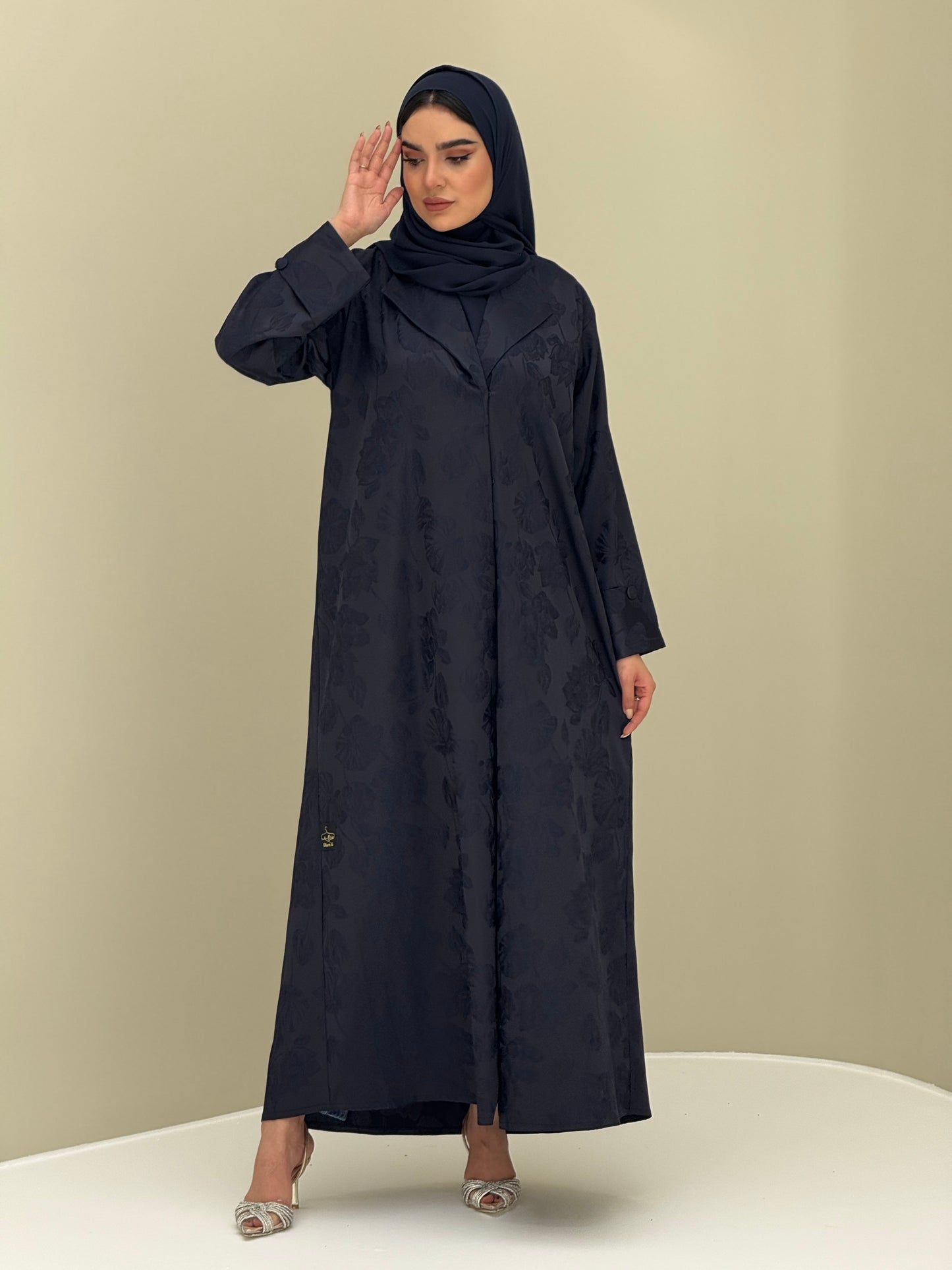 A woman wearing a navy blue abaya with a jacquard fabric, including a matching sheila headscarf, standing against a beige background.
