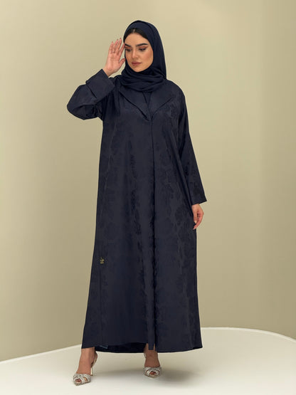 A woman wearing a navy blue abaya with a jacquard fabric, including a matching sheila headscarf, standing against a beige background.