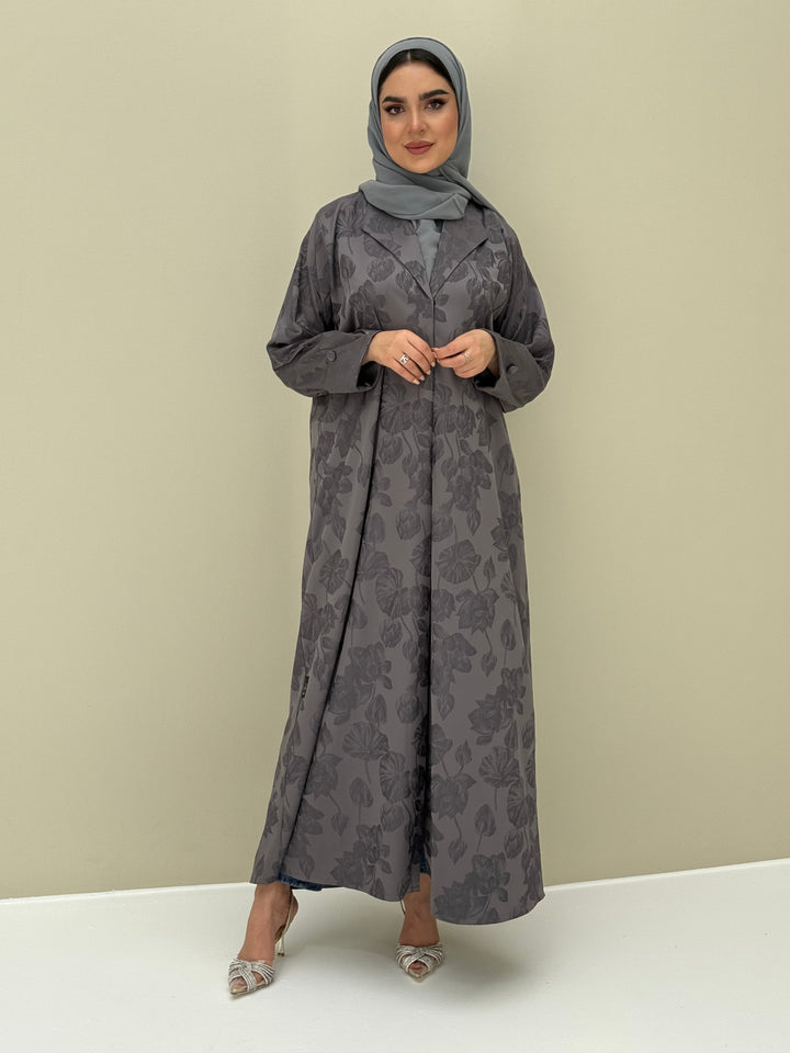 A woman wearing a steel grey abaya with a jacquard fabric, including a matching sheila headscarf, standing against a beige background.