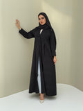 A woman standing in a neutral pose wearing a black Ice Cool Abaya flared coat style with lapel collar and a matching sheila, white shoes.
