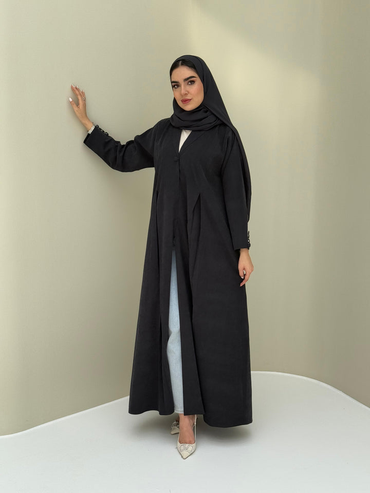 A woman standing in a neutral pose wearing a black Ice Cool Abaya flared coat style with lapel collar and a matching sheila, white shoes.