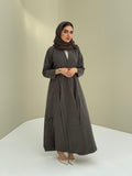 A woman standing in a neutral pose wearing a dark olive green Ice Cool Abaya flared coat style with lapel collar and a matching sheila, white shoes.