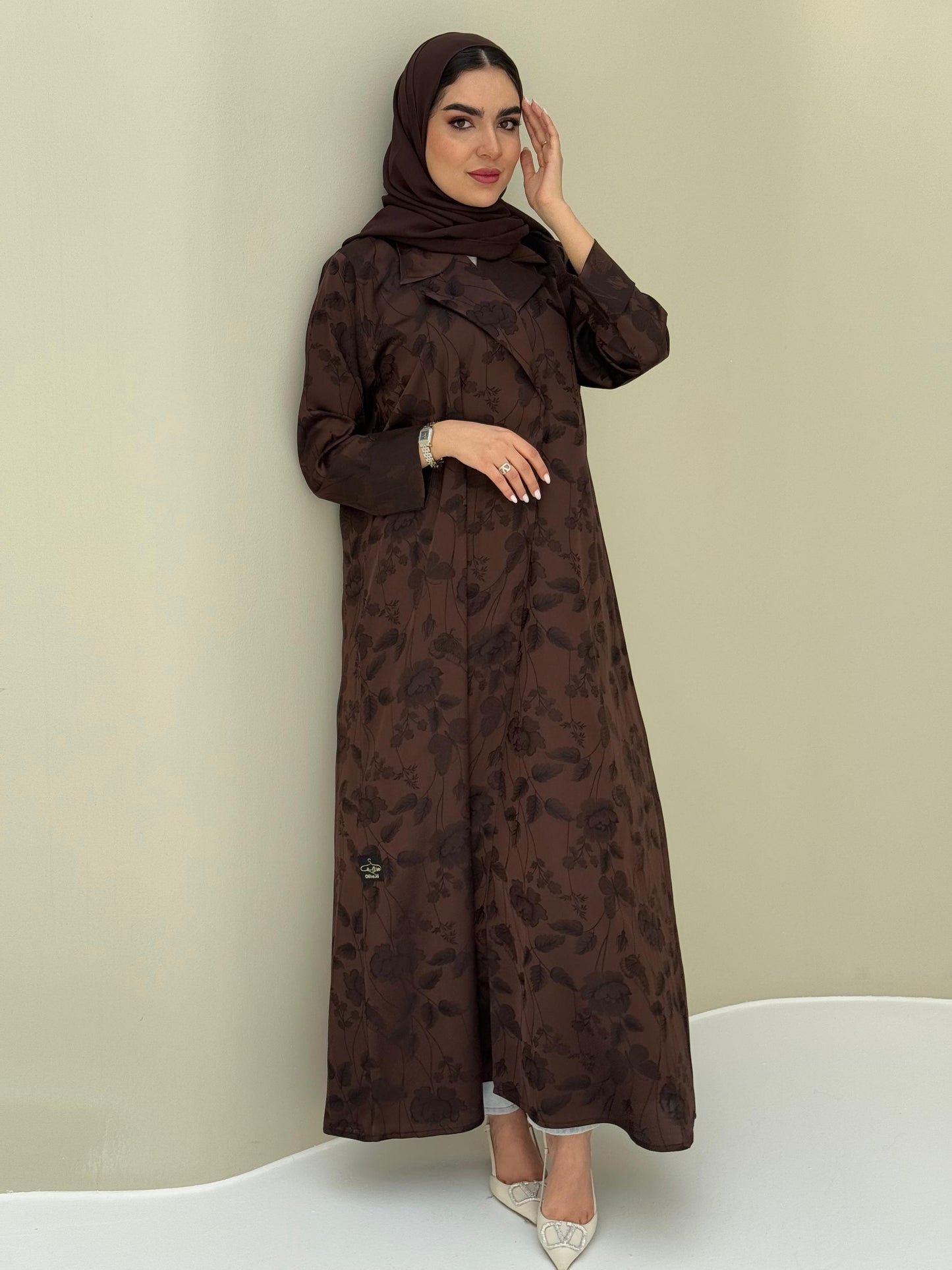 A woman wearing a dark brown abaya with a tonal floral print pattern and a matching sheila headscarf.