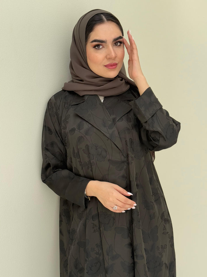 A woman wearing an olive green abaya with a tonal floral print pattern and a matching sheila headscarf.