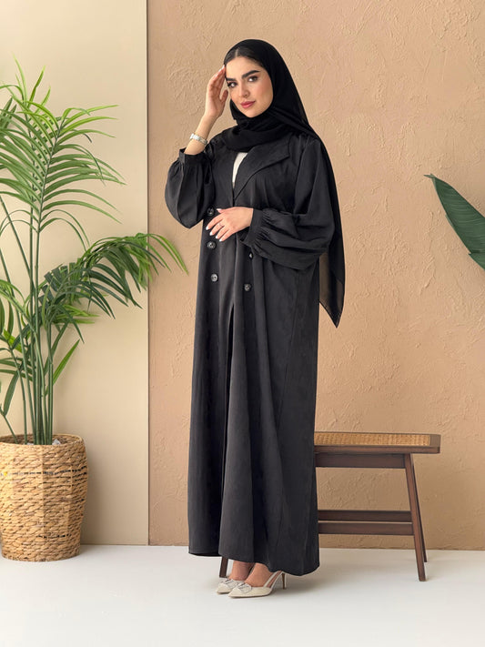 A woman standing in a room with a plant and a wooden chair in the background, wearing a black Abstract Satin abaya with Tonal brushstroke pattern, puffed cuffs, lapel collar, long sleeves and a matching Sheila headscarf
