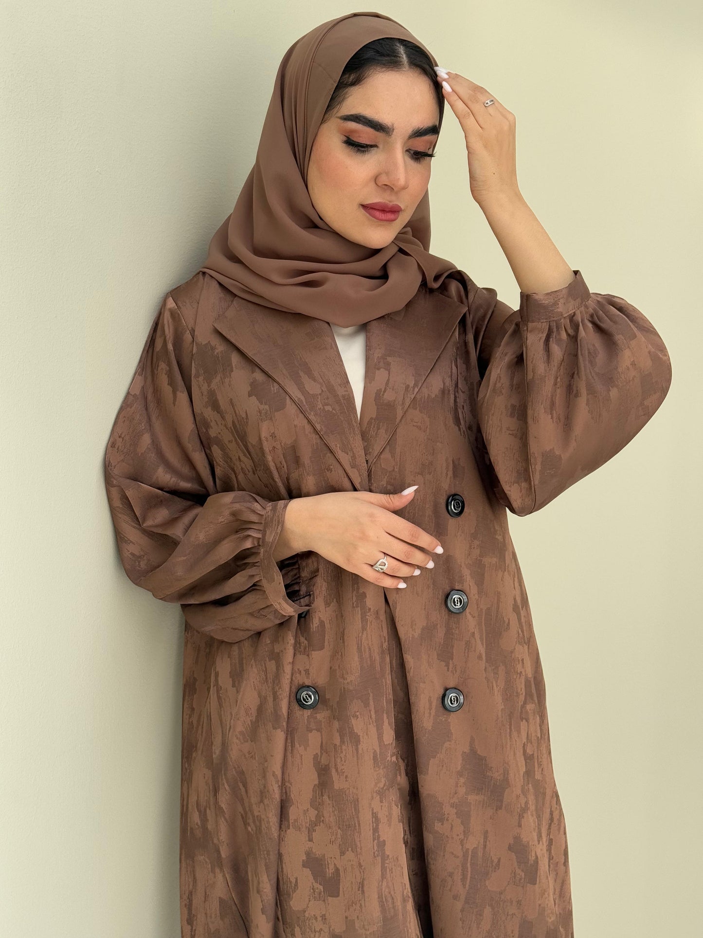A woman standing in a room with cream background, wearing a bronze brown Abstract Satin abaya with Tonal brushstroke pattern, puffed cuffs, lapel collar, long sleeves and a matching Sheila headscarf