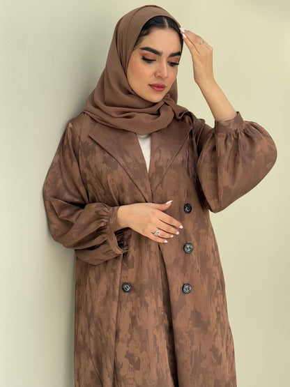 A woman standing in a room with cream background, wearing a bronze brown Abstract Satin abaya with Tonal brushstroke pattern, puffed cuffs, lapel collar, long sleeves and a matching Sheila headscarf