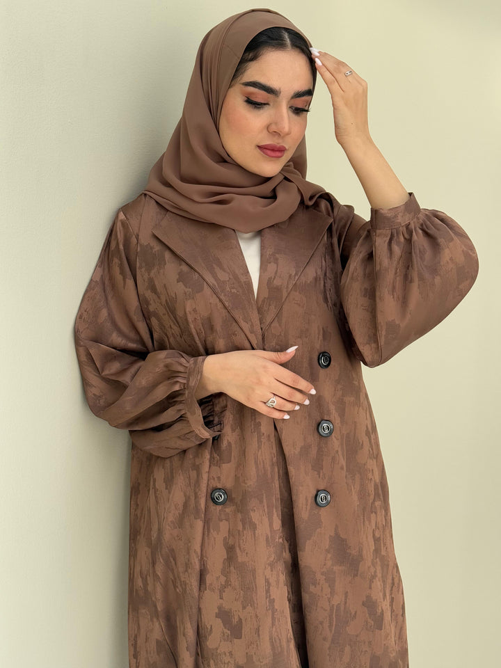 A woman standing in a room with cream background, wearing a bronze brown Abstract Satin abaya with Tonal brushstroke pattern, puffed cuffs, lapel collar, long sleeves and a matching Sheila headscarf