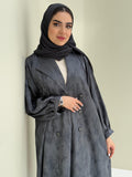 A woman standing in a room with cream background, wearing a dark grey Abstract Satin abaya with Tonal brushstroke pattern, puffed cuffs, lapel collar, long sleeves and a matching Sheila headscarf