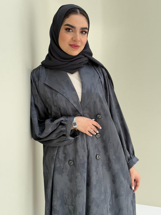 A woman standing in a room with cream background, wearing a dark grey Abstract Satin abaya with Tonal brushstroke pattern, puffed cuffs, lapel collar, long sleeves and a matching Sheila headscarf