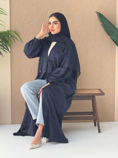 A woman sitting in a room with cream background, wearing a navy blue Abstract Satin abaya with Tonal brushstroke pattern, puffed cuffs, lapel collar, long sleeves and a matching Sheila headscarf