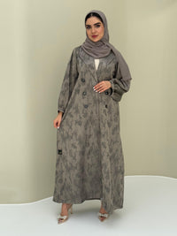 A woman standing in a room with a plant and a wooden chair in the background, wearing a sage green Abstract Satin abaya with Tonal brushstroke pattern, puffed cuffs, lapel collar, long sleeves and a matching Sheila headscarf