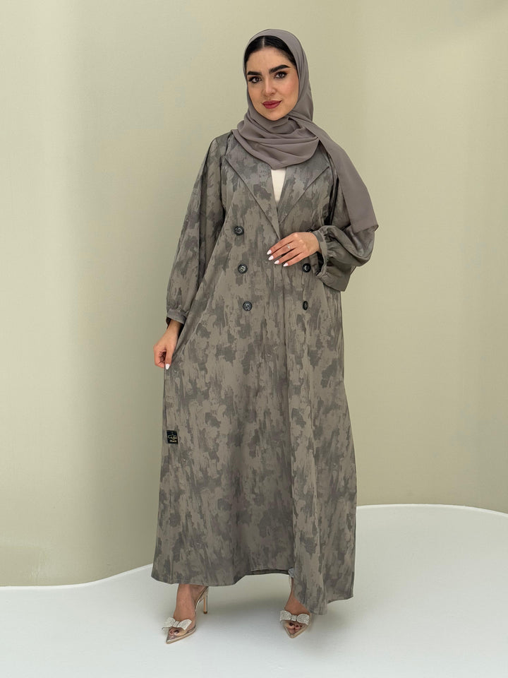 A woman standing in a room with a plant and a wooden chair in the background, wearing a sage green Abstract Satin abaya with Tonal brushstroke pattern, puffed cuffs, lapel collar, long sleeves and a matching Sheila headscarf