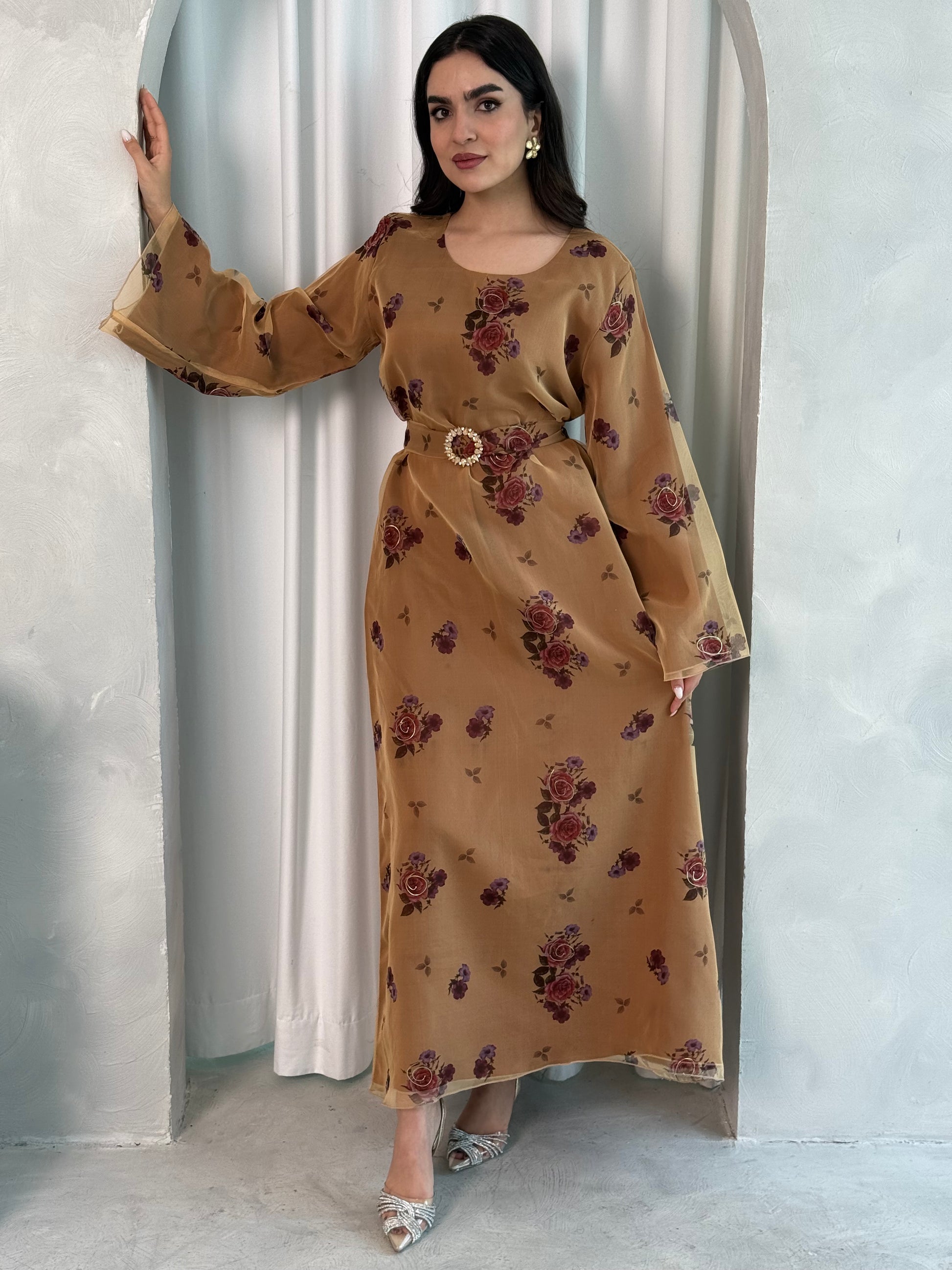 A woman wearing a Bronze & Burgundy printed organza (Lined) jalabiya dress with long sleeves and a belt, standing in front of a neutral backdrop.