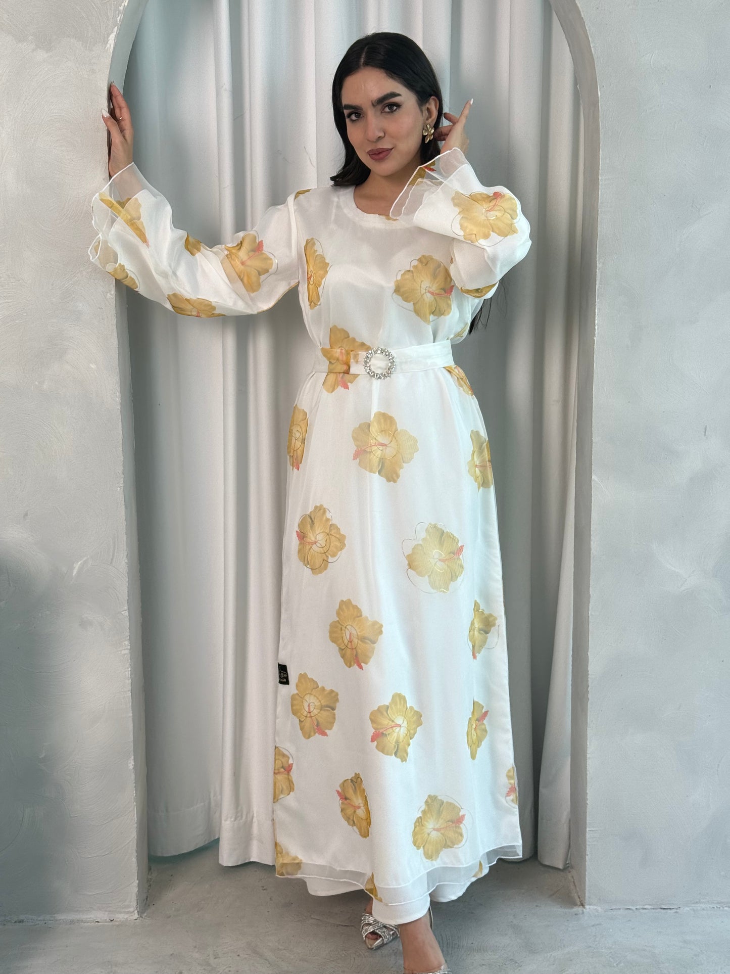 A woman wearing a White & Yellow  printed organza (Lined) jalabiya dress with long sleeves and a belt, standing in front of a neutral backdrop.