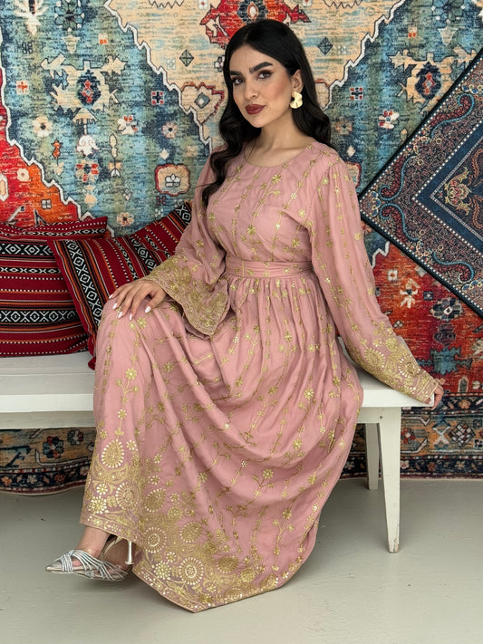A woman seated on a bench against a colorful, patterned backdrop, wearing a dusty pink A-line Jalabiya with wide sleeves with gold embroidered details and long sleeves.