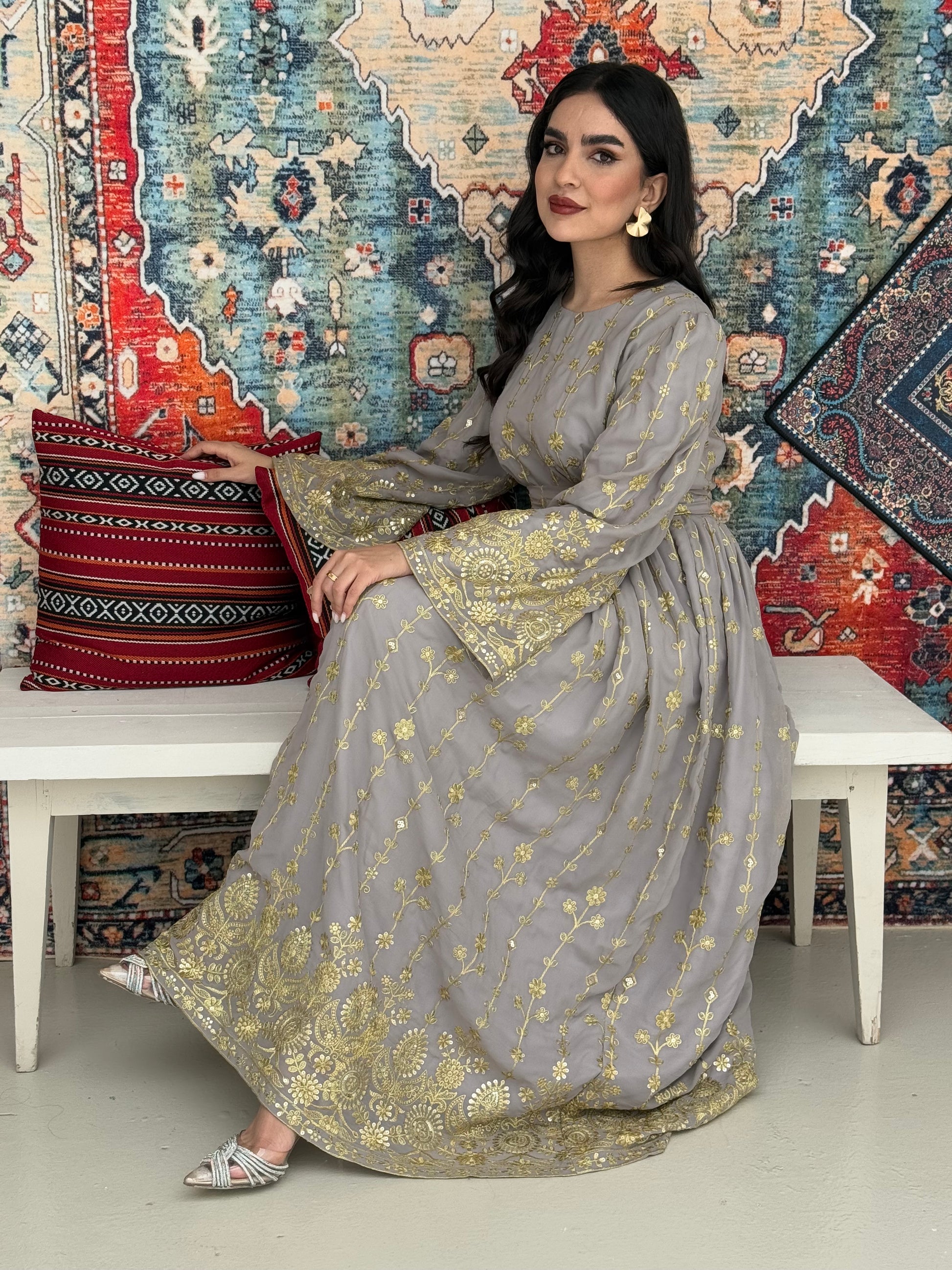 A woman seated on a bench against a colorful, patterned backdrop, wearing a grey A-line Jalabiya with wide sleeves with gold embroidered details and long sleeves.