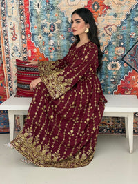 A woman seated on a bench against a colorful, patterned backdrop, wearing a maroon A-line Jalabiya with wide sleeves with gold embroidered details and long sleeves.
