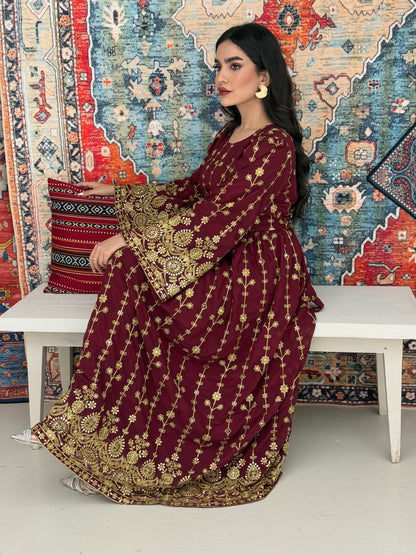 A woman seated on a bench against a colorful, patterned backdrop, wearing a maroon A-line Jalabiya with wide sleeves with gold embroidered details and long sleeves.