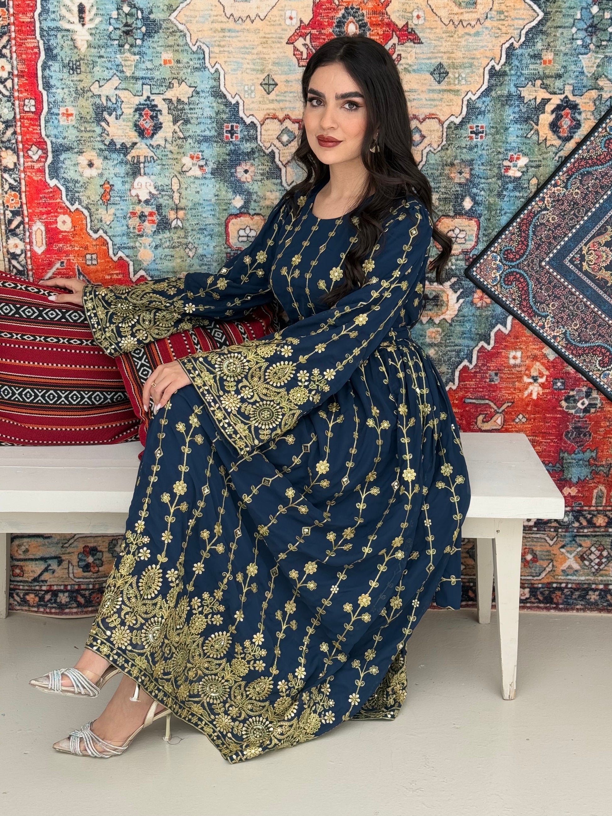 A woman seated on a bench against a colorful, patterned backdrop, wearing a Navy Blue A-line Jalabiya with wide sleeves with gold embroidered details and long sleeves.